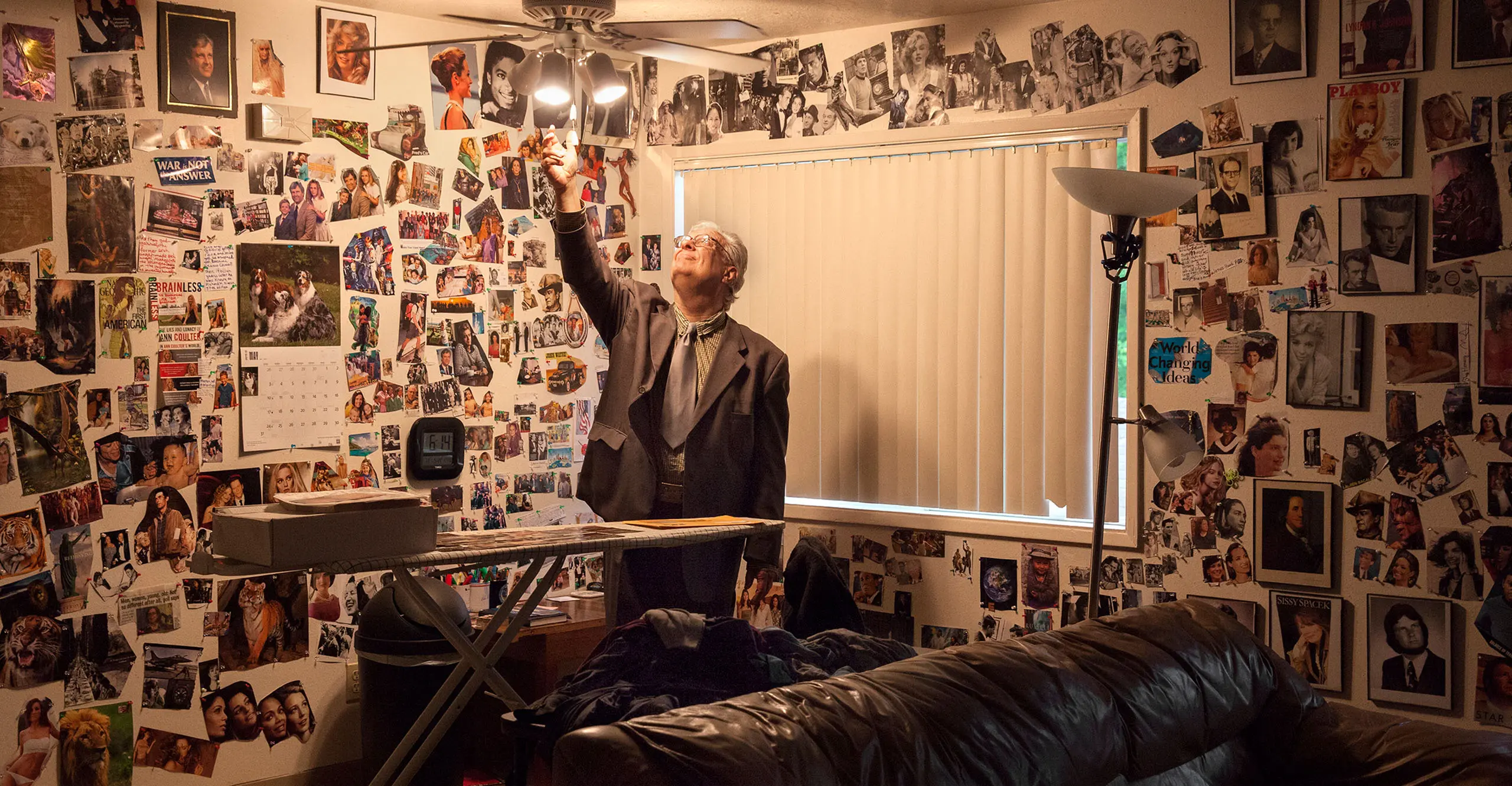 Image of an elderly man in his living room reaching up to the ceiling lamp as his walls are covered in magazine cutouts by bieke depoorter at the photographers gallery.